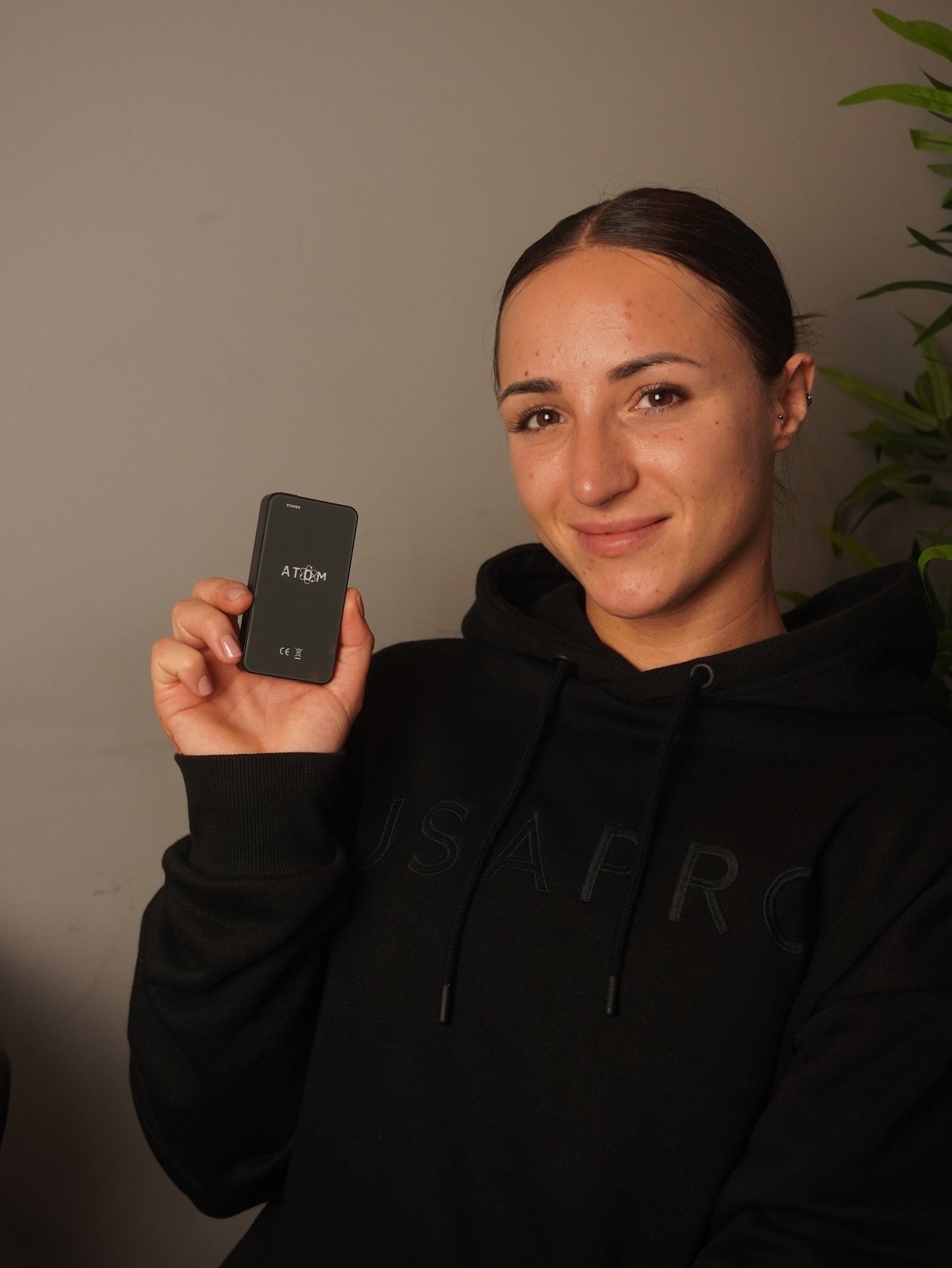 Woman in a black hoody holding a frequency device with a white wall in the background.